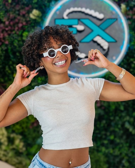Woman smiling with SmileFX clear aligners and sunglasses outdoors.