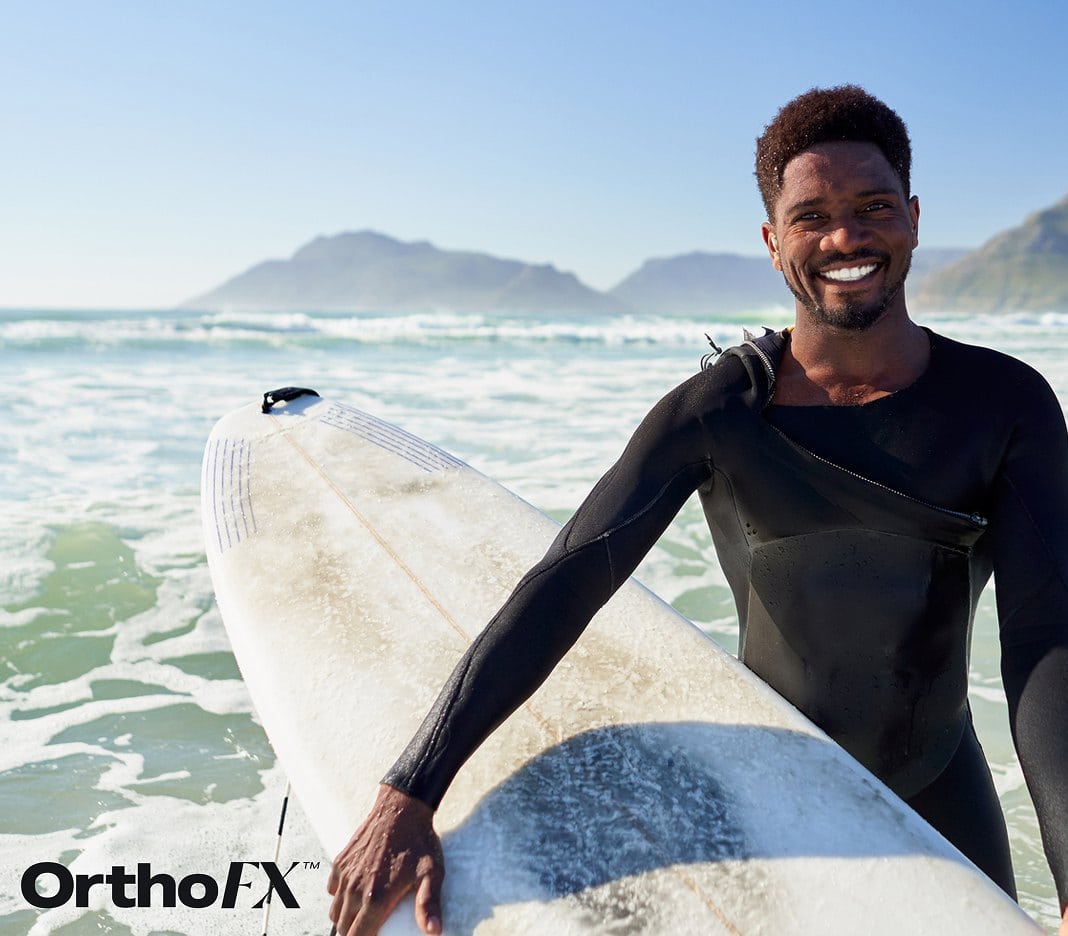 Smiling man with surfboard at the beach, promoting orthodontic products.